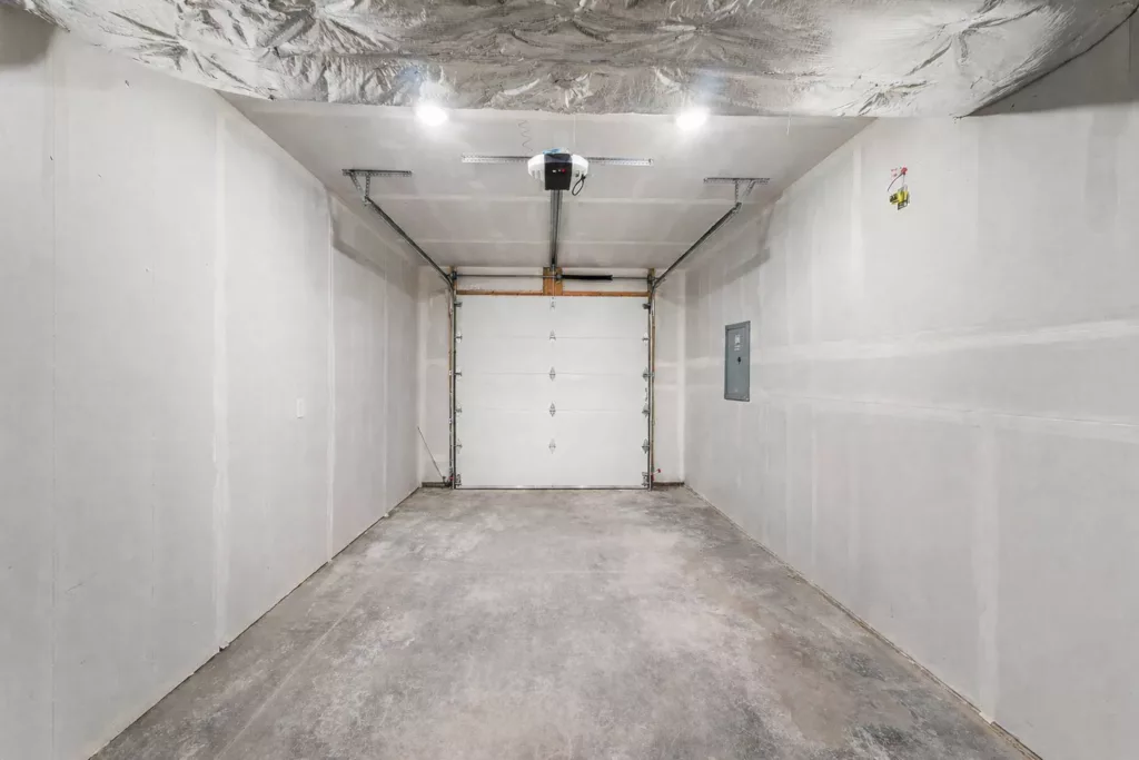 Empty garage with concrete floor, white walls, overhead door, ceiling lights, and an electrical panel on the right wall.